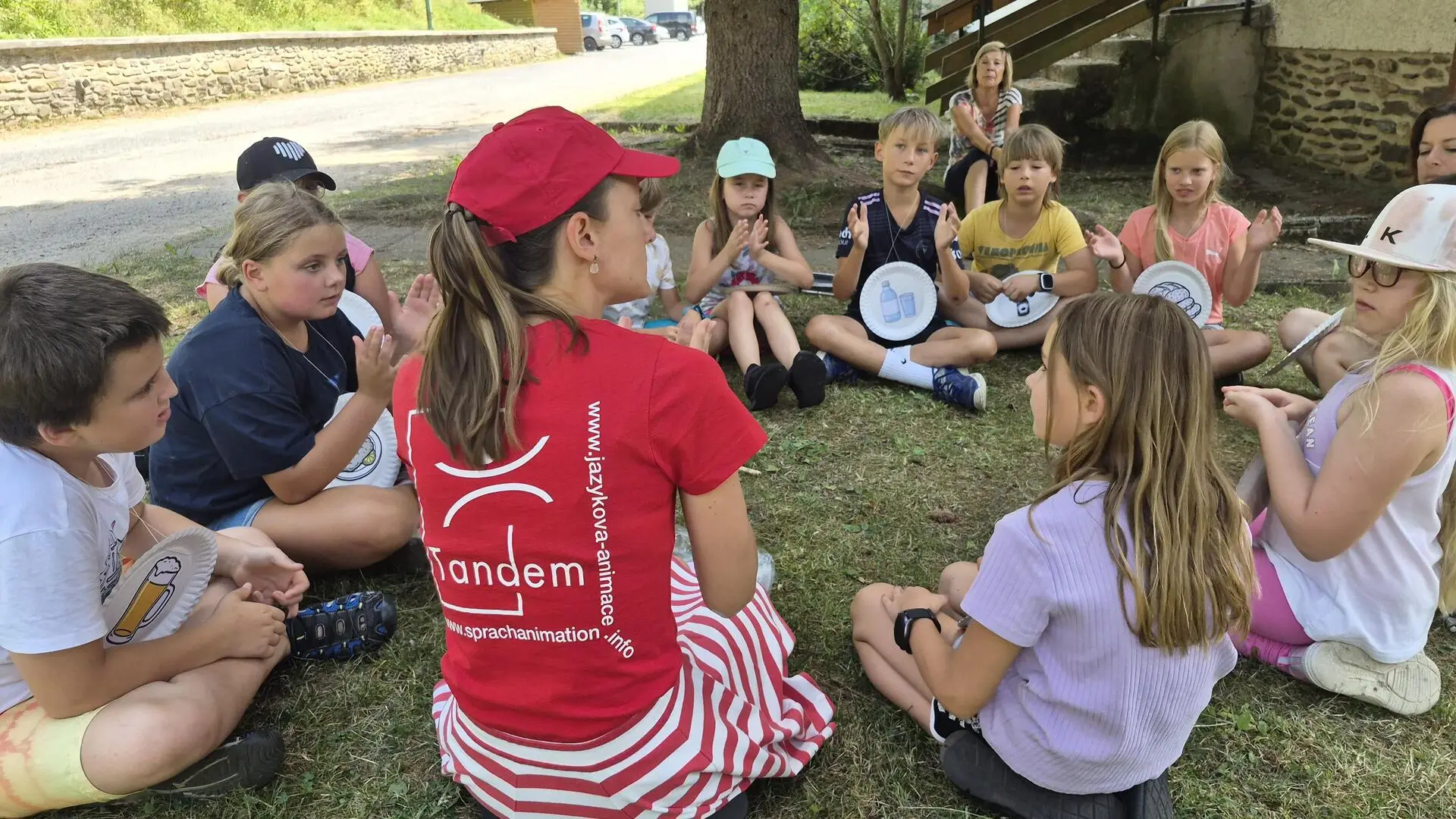 Ahoj-Hallo Sommercamp 2026 Eine Sprachanimateurin sitzt in einem Kreis mit Kindern